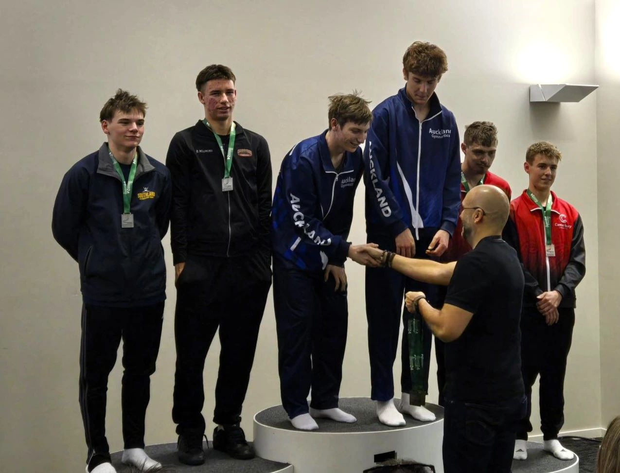 Group of six athletes standing on a podium. Christi Moller shaking hand with winner, handing him his medal. Athletes are proud and excited